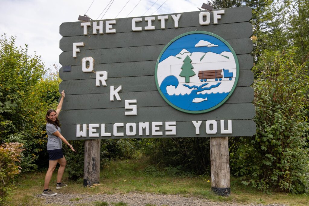 Ein großes hölzernes Schild mit der Aufschrift „THE CITY OF FORKS WELCOMES YOU“ und einem Emblem von Bergen und einem Lastwagen wird von einer Frau im Freien begleitet.